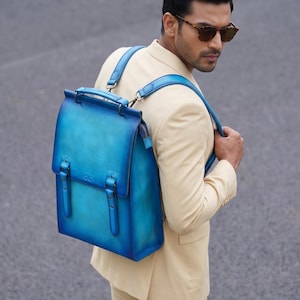 May include: A blue leather backpack with a gradient effect, featuring a top handle, adjustable straps, and buckle closures. The bag is carried by a person wearing a cream-colored suit.