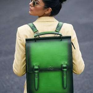 Pode incluir: Mochila de couro verde com fecho de aba, duas correias de fivela e uma alça superior. A mochila tem um efeito de cor gradiente e um pequeno design floral em relevo. As alças são verdes e presas na parte superior.