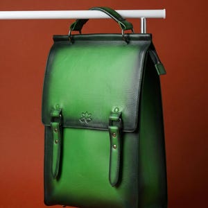 Pode incluir: Uma mochila de couro verde com fecho de aba e duas correias com fivela. A bolsa tem uma alça superior e um fecho de zíper. O couro tem um efeito gradiente, passando de um verde mais claro para um tom mais escuro. Um pequeno emblema é visível na frente.