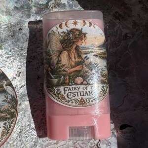 May include: A round metal tin and a pink plastic stick container, both featuring a circular label with an illustration of a fairy in a landscape and the text Fairy of the Estuary. The labels show a fairy with wings, flowers, and a bird.