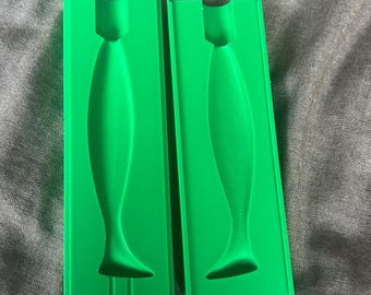 May include: Two bright green, rectangular molds with a fish-shaped cavity. The molds are made of a solid material and are designed to create fish-shaped objects. The molds are placed on a gray fabric surface.