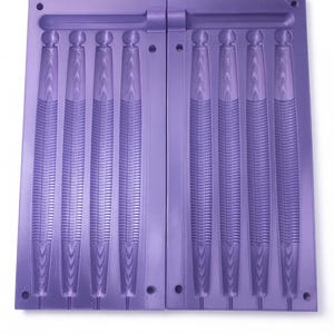 May include: A two-part, lavender-coloured mould for creating multiple identical objects. The mould features six cavities, each shaped like a stylized, elongated object with a textured surface. The mould is designed to be opened and closed, with a hinge on one side.