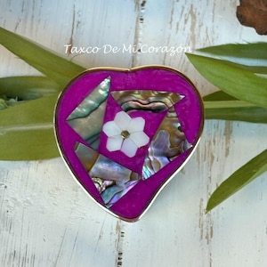 May include: A heart-shaped jewelry box with a vibrant magenta interior and a silver-colored border. The box is adorned with iridescent abalone shell fragments and a white flower. The text "Taxco De Mi Corazón" is visible.