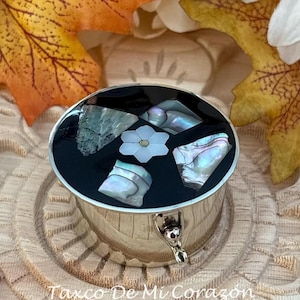 May include: A silver-colored, oval-shaped trinket box with a black lid. The lid is inlaid with iridescent shell fragments and a white flower. The text "Taxco De Mi Corazon" is printed on the base.