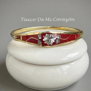 Könnte beinhalten: Ein goldfarbenes Klapparmband mit einer zentralen roten Einlage, die mit einem floralen Perlmutt-Mosaik-Design verziert ist. Der Text Taxco De Mi Corazón ist über dem Armband sichtbar. Das Schmuckstück hat eine polierte Metalloberfläche.