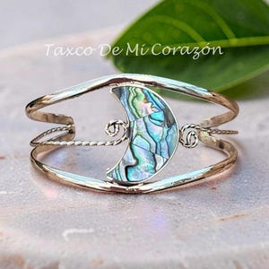 Peut inclure: Un bracelet manchette argenté avec une incrustation centrale en forme de croissant de lune en nacre irisée. La bande métallique présente un design fendu avec des accents de fil torsadé de chaque côté. Le texte Taxco De Mi Corazón est visible.