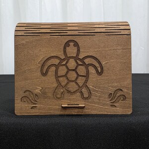以下が含まれることがあります： 彫刻されたカメのデザインが施された長方形の木箱。箱は湾曲した蓋と小さな引き出しが付いています。カメが中心のデザインで、下に波模様があります。木材は暖かく茶色で、収納に最適です。