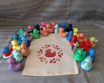 Puede incluir: Una colección de patitos de goma de colores en varios tonos de rosa, naranja, verde, azul, amarillo y negro. Una bolsa de lona con un gráfico de pato está en el centro. Los patos están dispuestos alrededor de la bolsa.