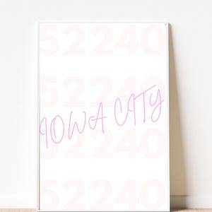 Może przedstawiać: Oprawiony biały nadruk z napisem "IOWA CITY" w różowej, odręcznej czcionce. Tło przedstawia wyblakły wzór numeru "52240" w kolorze różowym. Nadruk jest prezentowany na jasnej drewnianej podłodze.
