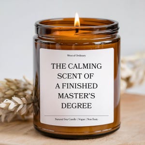 Puede incluir: Una vela encendida de vidrio ámbar con una etiqueta blanca. La etiqueta dice "THE CALMING SCENT OF A FINISHED MASTER'S DEGREE". La vela está etiquetada como "West of Ordinary" y está hecha de soja natural, vegana y no tóxica.