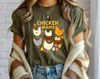 May include: Olive green t-shirt with the text "CHICKEN MAMA" and a graphic of several chickens, nests, and eggs. The design features various chicken breeds in white, orange, and gray, with colorful eggs and floral accents. The shirt has rolled-up sleeves.