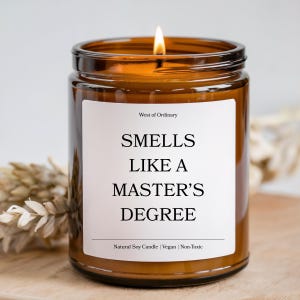 May include: A lit brown glass jar candle with a white label. The label reads "SMELLS LIKE A MASTER'S DEGREE" in black text. The candle is labeled "West of Ordinary" and is made of natural soy, vegan, and non-toxic materials.