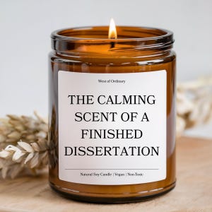 May include: A lit brown glass jar candle with a white label. The label reads "THE CALMING SCENT OF A FINISHED DISSERTATION". The candle is labeled "West of Ordinary" and "Natural Soy Candle | Vegan | Non-Toxic".