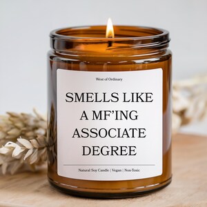 May include: A lit, brown glass jar candle with a white label. The label reads "SMELLS LIKE A MF'ING ASSOCIATE DEGREE." The candle is labeled "West of Ordinary" and is made of natural soy, vegan, and non-toxic materials.