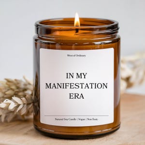 Peut inclure: Une bougie en cire de soja allumée dans un pot en verre brun. L'étiquette blanche indique "IN MY MANIFESTATION ERA", avec "West of Ordinary" au-dessus et "Natural Soy Candle | Vegan | Non-Toxic" en dessous. La bougie est sur une surface en bois.