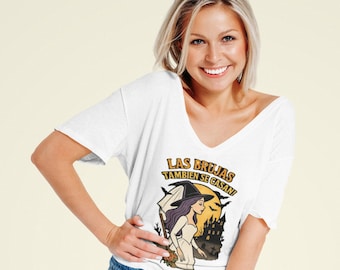 Könnte beinhalten: Weißes V-Ausschnitt-T-Shirt mit einer Grafik einer Hexe in einem Hochzeitskleid und dem Text "LAS BRUJAS TAMBIEN SE CASAN!" in Gelb und Schwarz. Das Design beinhaltet eine Silhouette eines Spukhauses und Fledermäuse.