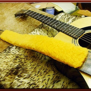 May include: A yellow guitar strap made of soft, fluffy material. The strap is lying on a brown, fuzzy surface and is attached to a brown acoustic guitar.