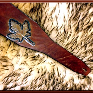 May include: A brown leather guitar strap with a tooled leaf design. The strap is lying on a white and brown fur surface.