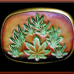 Puede incluir: Hebilla de cinturón rectangular con borde dorado. La hebilla presenta un fondo marrón y naranja con hojas verdes de cannabis. Las hojas están intrincadamente detalladas y dispuestas en un patrón simétrico.