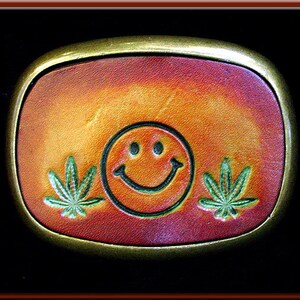 Op de afbeelding: Een leren riemgesp met een smiley-gezicht en groene marihuana-bladontwerpen op een rood, oranje en gele achtergrond.