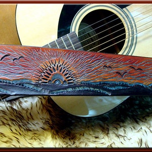Peut inclure: Une sangle de guitare en cuir marron avec un motif ciselé d'un coucher de soleil avec des oiseaux volant au-dessus d'une étendue d'eau. La sangle est posée sur une guitare acoustique.