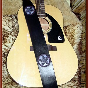 May include: A black leather guitar strap with two silver pentagram designs. The strap is on a light brown acoustic guitar with a black pickguard.