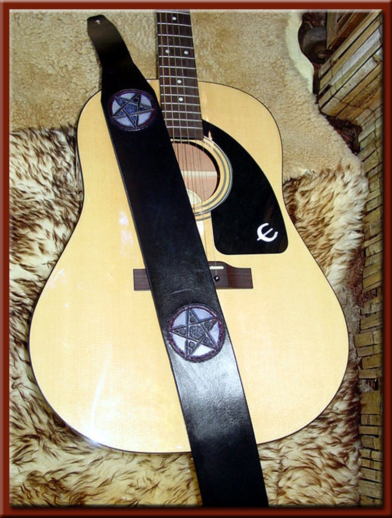 May include: A black leather guitar strap with two silver pentagram designs. The strap is on a light brown acoustic guitar with a black pickguard.