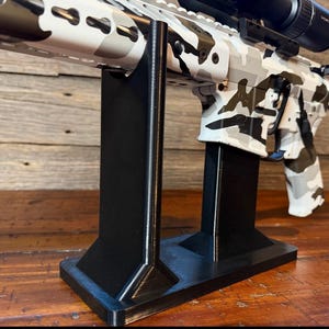 May include: A black plastic display stand with two vertical supports designed to hold a rifle. The rifle features a white, gray, and black camouflage pattern across the receiver, handguard, and stock, and is equipped with a black Vortex brand scope.