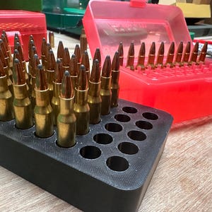 May include: A close-up shot of ammunition cartridges in a black holder and a red plastic case. The cartridges have brass casings and pointed tips. The black holder has multiple circular holes to hold the cartridges upright.
