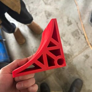 May include: A bright red, triangular-shaped wheel chock featuring a hollow, geometric lattice structure with multiple support struts and a circular opening at the base corner, designed for securing a vehicle or trailer wheel in place.
