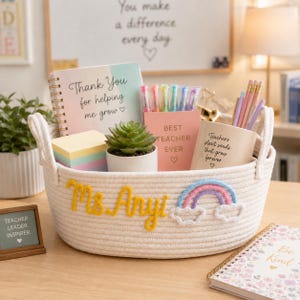 Può includere: Un cesto intrecciato color crema pieno di regali per insegnanti. Include un taccuino, penne, matite, una succulenta e un blocco note. Il cesto è decorato con un arcobaleno e il nome "Ms. Amy" in filo giallo.