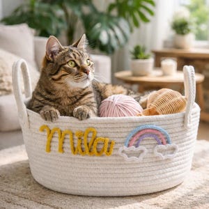 Puede incluir: Un gato atigrado descansa en una cesta de cuerda blanca con asas. La cesta está decorada con el nombre "Mila" en hilo amarillo y un aplique de arcoíris. Dentro de la cesta hay lana rosa y un objeto tejido.