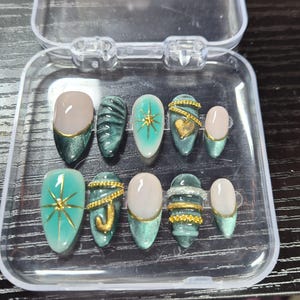 May include: A set of ten artificial nails in a clear plastic case. The nails feature various designs, including teal and gold accents, gold starbursts, and gold heart details. Some nails have a nude-colored base with gold tips.