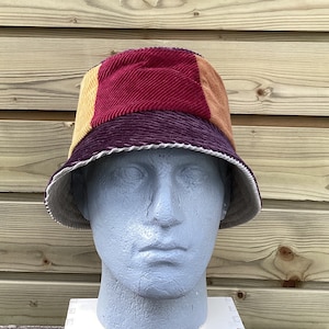 May include: A multi-colored corduroy bucket hat with a burgundy, tan, and purple color scheme. The hat is on a mannequin head.