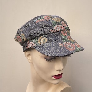 May include: A grey newsboy cap featuring a floral design in pink, yellow, and green. The cap has a short peak and a structured crown. The fabric has a subtle sheen, and the floral design is intricately woven into the material.