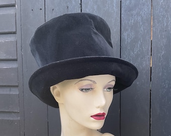 May include: A black top hat with a wide brim. The hat is made of a soft, velvety material. The hat is displayed on a mannequin head with painted features and red lipstick. The background is a dark wooden fence.
