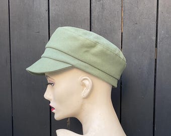 May include: A light green, textured newsboy cap with a short brim. The cap is displayed on a mannequin head against a dark wooden background. The cap's design features a flat top and a structured shape, suitable for various styles.