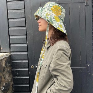 K&ouml;nnte beinhalten: Ein Eimerhut mit floralem Muster mit gelben und gr&uuml;nen Blumen auf hellem Hintergrund. Der Hut hat eine breite Krempe und B&auml;nder, die herunterh&auml;ngen. Die Person tr&auml;gt eine gr&uuml;ne Jacke.