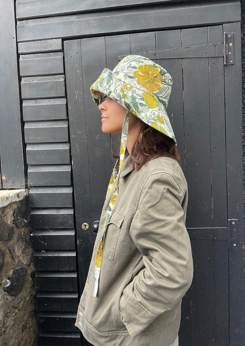 K&ouml;nnte beinhalten: Ein Eimerhut mit floralem Muster mit gelben und gr&uuml;nen Blumen auf hellem Hintergrund. Der Hut hat eine breite Krempe und B&auml;nder, die herunterh&auml;ngen. Die Person tr&auml;gt eine gr&uuml;ne Jacke.