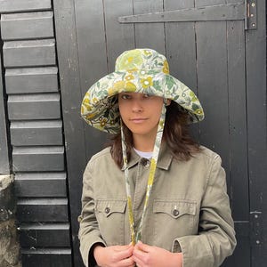 K&ouml;nnte beinhalten: Ein Sonnenhut mit breiter Krempe und einem Blumenmuster in Gelb- und Gr&uuml;nt&ouml;nen. Der Hut hat lange B&auml;nder und wird mit einer gr&uuml;nen Jacke getragen. Der Hintergrund ist eine dunkle Holzt&uuml;r.