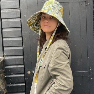 K&ouml;nnte beinhalten: Ein breitkrempiger Sonnenhut mit einem Blumenmuster in Gelb, Gr&uuml;n und Wei&szlig;. Der Hut hat einen passenden Kinnriemen. Die Person tr&auml;gt eine hell olivgr&uuml;ne Jacke.
