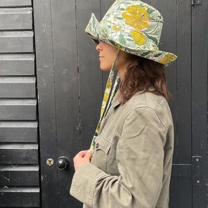 K&ouml;nnte beinhalten: Ein breitkrempiger Hut mit einem gelb-gr&uuml;nen Blumenmuster und einem passenden Stoffband. Der Hut wird mit einer gr&uuml;nen Jacke getragen. Der Hintergrund ist eine schwarze Holzt&uuml;r.