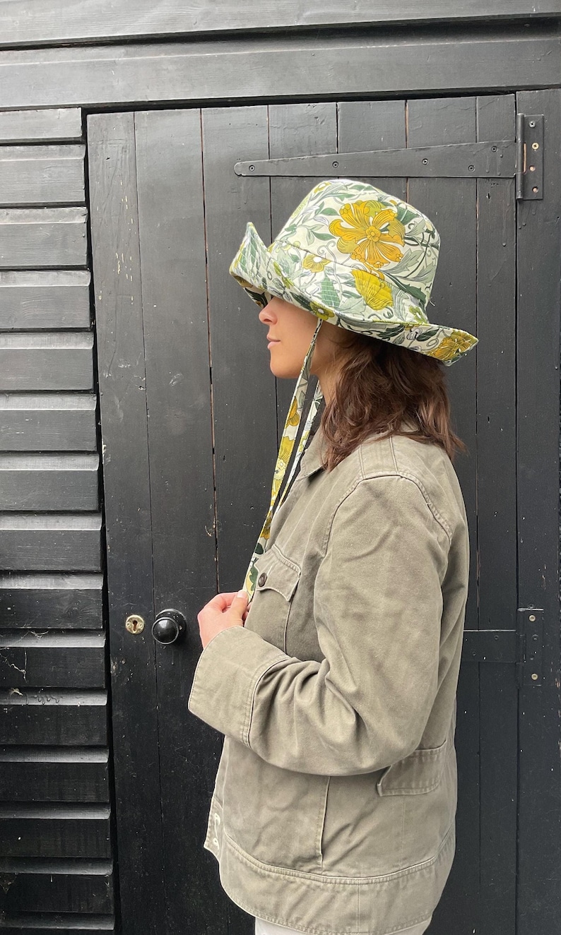 K&ouml;nnte beinhalten: Ein breitkrempiger Hut mit einem gelb-gr&uuml;nen Blumenmuster und einem passenden Stoffband. Der Hut wird mit einer gr&uuml;nen Jacke getragen. Der Hintergrund ist eine schwarze Holzt&uuml;r.