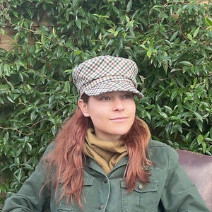 May include: A brown, green, and white checked newsboy cap. The cap is worn by a person with auburn hair, a green jacket, and a tan scarf. The background is a dense wall of green leaves.