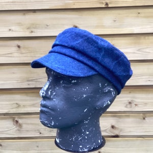 Puede incluir: Una gorra de newsboy de mezclilla azul con una visera doblada. La gorra está en una cabeza de maniquí negra.