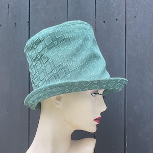 May include: A teal green top hat with a diamond pattern. The hat has a wide brim and a tall, cylindrical crown. The hat is displayed on a mannequin head.