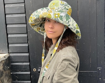 May include: A wide-brimmed hat with a floral pattern in shades of green, yellow, and white. The hat has a rounded crown and a wide, floppy brim. The hat is tied with matching fabric straps. The person is wearing an olive green jacket.