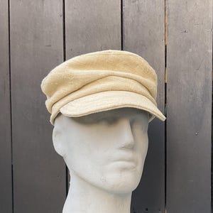 May include: A beige newsboy cap with a short brim. The cap is made of a textured fabric and sits atop a white mannequin head. The background is a dark gray wooden surface.