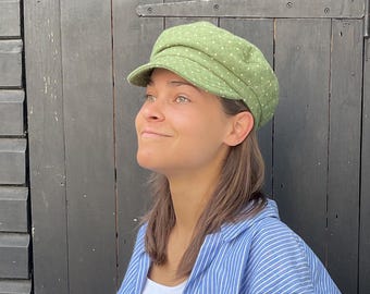 Könnte beinhalten: Hellgrüne Schiebermütze mit weißen Tupfen. Die Mütze hat einen kurzen Schirm und eine runde Krone. Die Person trägt ein blau-weiß gestreiftes Hemd.