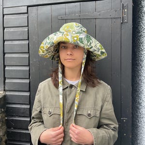 K&ouml;nnte beinhalten: Ein breitkrempiger Hut mit einem Blumenmuster in Gr&uuml;n-, Gelb- und Wei&szlig;t&ouml;nen. Der Hut hat lange B&auml;nder. Die Person im Bild tr&auml;gt eine olivgr&uuml;ne Jacke.